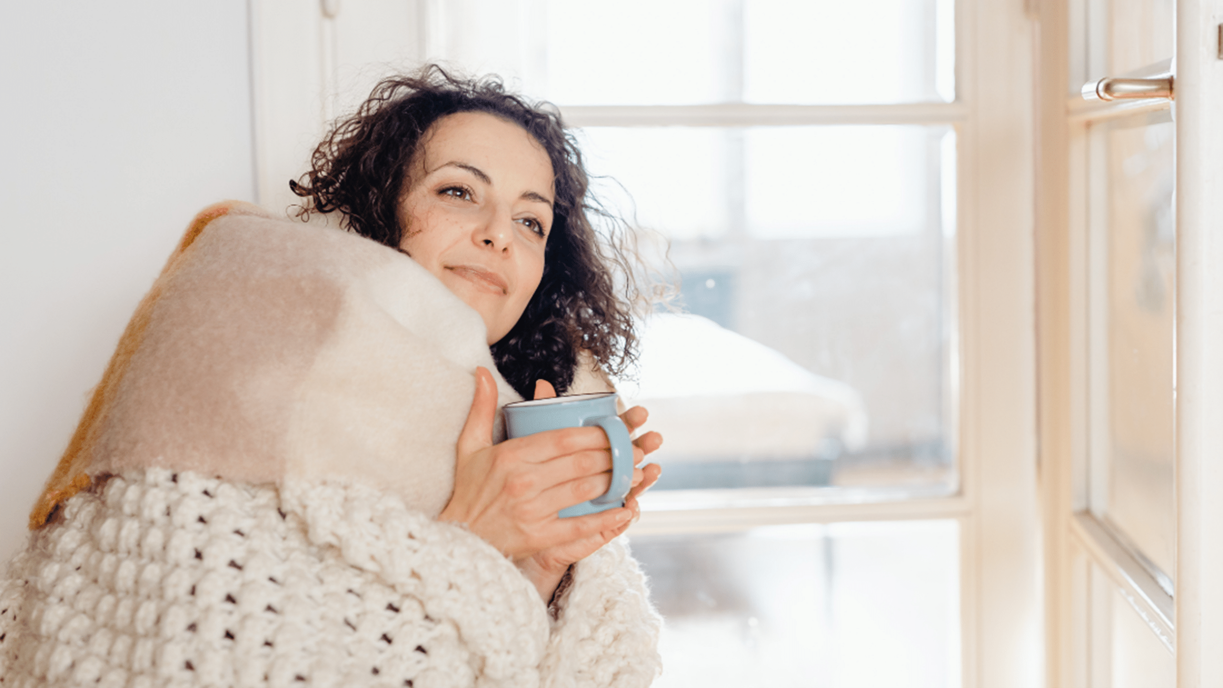Woman snuggled next to window in blanket holding mug